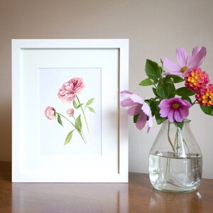 May include: A white frame containing a watercolour painting of pink peonies. A vase with pink and orange flowers sits on a wooden table.