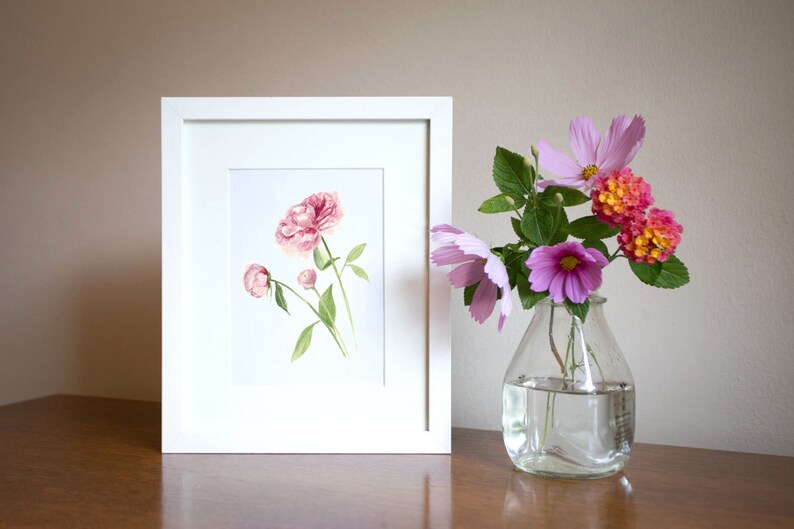 May include: A white frame containing a watercolour painting of pink peonies. A vase with pink and orange flowers sits on a wooden table.