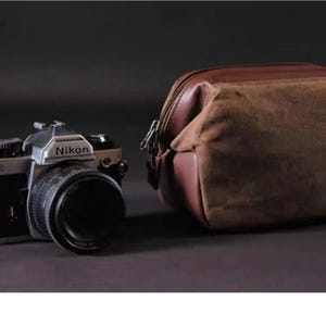 Może przedstawiać: Vintage aparat fotograficzny Nikon F2 SLR z czarnym obiektywem i srebrnymi akcentami, obok brązowej torby na aparat z płótna i skóry. Aparat i torba są na ciemnym tle.