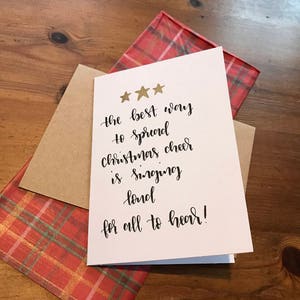 以下が含まれることがあります： 金色の星と手書きのテキストが書かれた白いクリスマスカード。「クリスマスを広める最良の方法は、みんなが聞けるように大声で歌うことです！」 カードは、赤いチェック柄の封筒と茶色の封筒の上に置かれています。