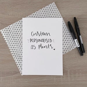 Op de afbeelding: Een witte A5-afdruk met de woorden "Custom PERSONALISED A5 prints" in zwarte kalligrafie. De afdruk ligt op een papier met stippen en wordt vergezeld door twee zwarte markers op een houten oppervlak.