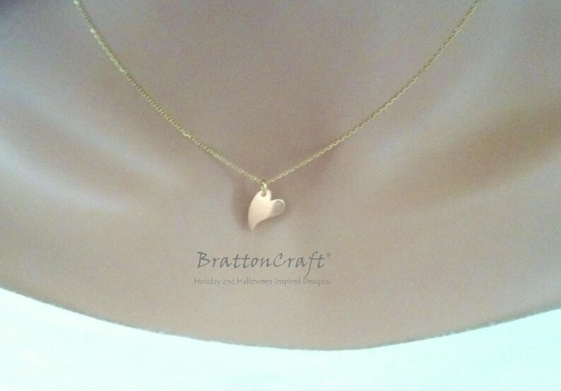May include: A gold chain necklace with a small, flat, gold heart pendant. The necklace is on a model's neck.
