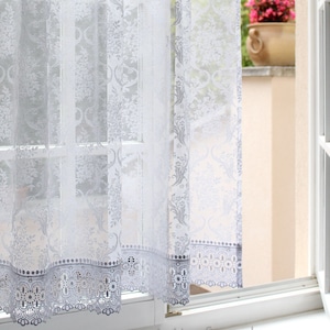 Op de afbeelding: Witte kantgordijnen met een delicaat bloemenpatroon en een gegolfde zoom. De gordijnen hangen in een raam met uitzicht op een tuin.
