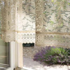 Könnte beinhalten: Zwei durchsichtige weiße Gardinen aus Spitze mit einem Blumenmuster und einem gewellten Saum. Die Gardinen hängen an einem Fenster und die Sonne scheint durch sie.