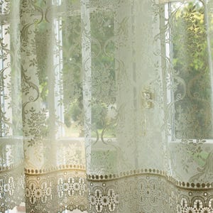 Op de afbeelding: Witte kantgordijnen met een delicaat bloemenpatroon en een gegolfde zoom. De gordijnen hangen aan een raam met uitzicht op groen gebladerte.