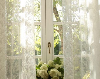 May include: Sheer white curtains featuring a delicate, repeating floral damask pattern hang in front of a window. The translucent fabric allows light to filter through, highlighting the intricate botanical details woven into the textile design.