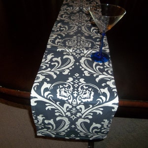 May include: A black and white damask table runner with a white background and black floral pattern. The runner is on a dark wood table with a clear glass on top.