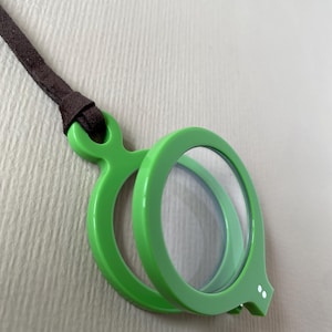 May include: A bright green magnifying glass pendant with a brown leather cord. The pendant has a circular lens and a rounded frame. The design includes a small, decorative element with two white dots.