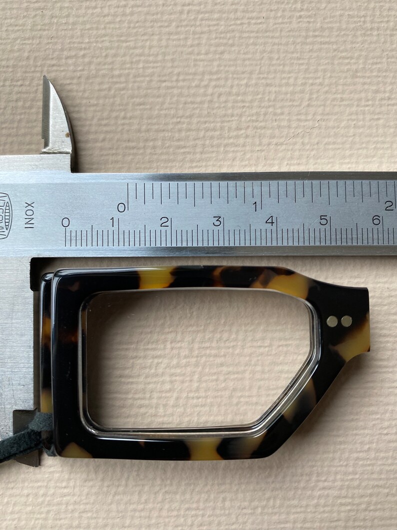 May include: A close-up of a tortoiseshell eyeglasses frame being measured with a ruler. The ruler shows the frame is approximately 5.5 cm wide.