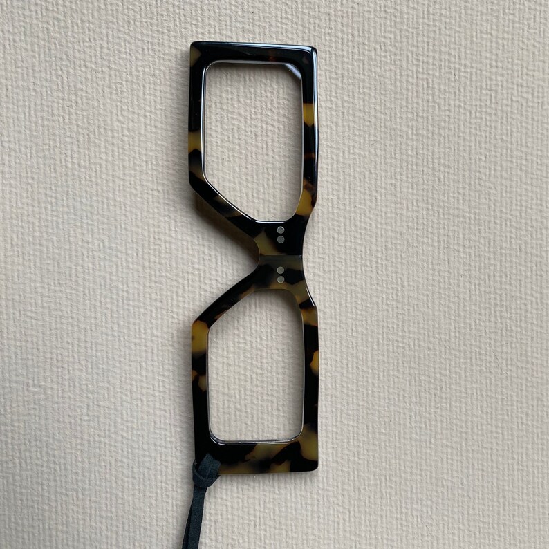 May include: Tortoise shell reading glasses with a black cord. The glasses have a unique geometric shape.