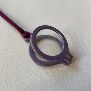 Puede incluir: Una lupa plegable de color morado con un cordón burdeos. La lupa tiene dos puntos dorados en el lateral.