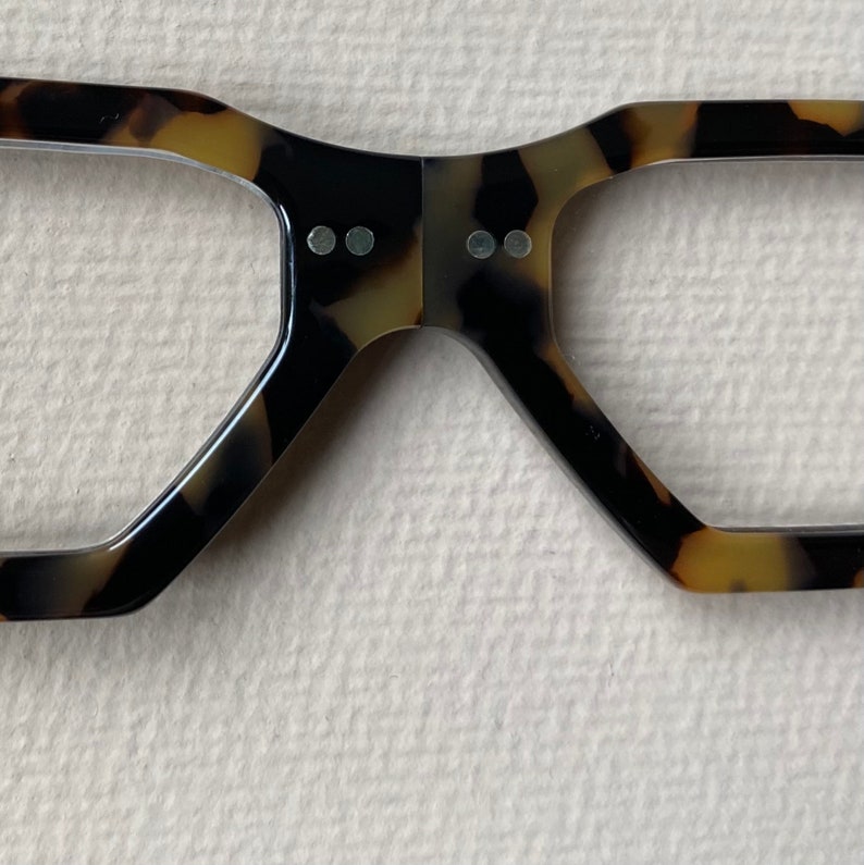 May include: A pair of tortoiseshell eyeglasses with a unique, angular frame design. The frame has a dark brown and light brown pattern.