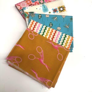 May include: A stack of four folded fabric squares. The top piece is mustard yellow with pink stork-shaped scissors. Below is a white strip with colorful wavy lines, a blue piece with small bird and figure motifs, and a pink patterned piece.