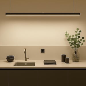 May include: A modern kitchen interior featuring a black linear pendant light above a sink and countertop. The countertop has a faucet, a sink, and a few dark bowls. A vase with green leaves and two dark cylinders are also on the countertop.
