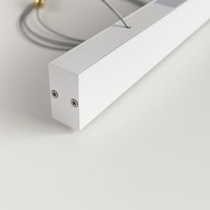 May include: A white rectangular LED light fixture with a recessed light source. The fixture is suspended by a thin, silver cable. Two screws are visible on the end of the fixture. The background is a plain white surface.