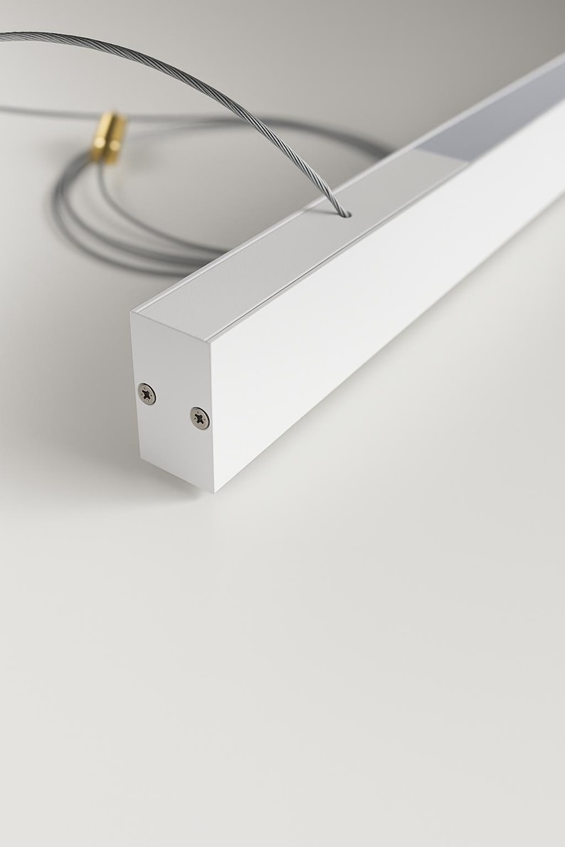 May include: A white rectangular LED light fixture with a recessed light source. The fixture is suspended by a thin, silver cable. Two screws are visible on the end of the fixture. The background is a plain white surface.
