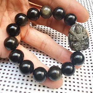 Può includere: Un braccialetto di perline nere e dorate con un ciondolo dorato raffigurante un Buddha. Il braccialetto è fatto di perline rotonde con una finitura lucida.