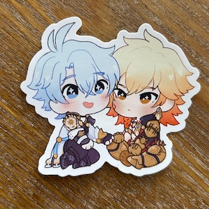 以下が含まれることがあります： 2人のちびスタイルのアニメキャラクターが描かれたダイカットステッカー。一人は水色の髪、もう一人はオレンジ色のアクセントが付いたブロンドの髪をしています。どちらも大きな目を持ち、笑顔です。ステッカーは木製の表面にあります。