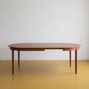 Pode incluir: Uma mesa de jantar oval de madeira com acabamento castanho quente, apresentando uma superfície lisa e quatro pernas cilíndricas cónicas. O design da mesa inclui uma costura visível no centro, indicando um mecanismo extensível para a peça.