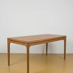 Op de afbeelding: Een rechthoekige houten tafel met een lichtbruine afwerking. De tafel heeft vier slanke, licht gebogen poten. Het tafelblad is glad en rechthoekig. De tafel staat tegen een witte muur en een geelbruine vloer.