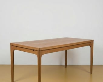 Può includere: Un tavolo rettangolare in legno con finitura marrone chiaro. Ha quattro gambe sottili e leggermente curve. Il piano del tavolo è liscio e rettangolare. Il tavolo è posto contro un muro bianco e un pavimento giallo-marrone.