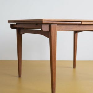 Peut inclure: Une table à manger rectangulaire en bois avec une finition brun chaud et un grain de bois visible. La table présente quatre pieds fuselés et une surface supérieure plate et lisse avec une jointure visible indiquant une conception extensible.