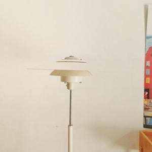 Puede incluir: Una lámpara de pie de color crema con un diseño de pantalla escalonada. La lámpara tiene un poste delgado de color blanquecino y una base circular. La pantalla se compone de varios elementos en forma de disco superpuestos. Diseño moderno de mediados de siglo.