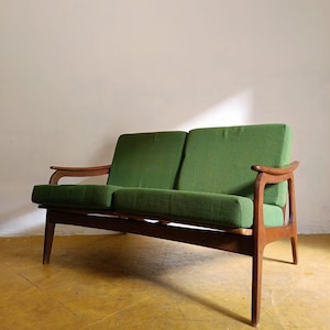 Op de afbeelding: Een tweezitsbank in een mid-century modern stijl. Het heeft een houten frame en poten, met groene kussens. Het ontwerp is minimalistisch, met gebogen armleuningen. Het lage profiel en de warme houttinten geven het een klassieke uitstraling.