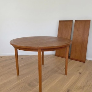 Pode incluir: Uma mesa de jantar redonda de madeira castanho-média com quatro pernas retas e cónicas. Duas extensões retangulares de madeira a condizer estão posicionadas na vertical atrás da mesa, exibindo um padrão de grão consistente e um acabamento quente.
