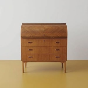 Op de afbeelding: Een houten rolltopbureau met een gebogen rolluikdeur met een visgraatpatroon in de houtnerf. De kast bevat drie horizontale lades onder het rolluik, elk voorzien van twee verzonken ovale handgrepen, rustend op vier taps toelopende poten.
