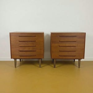 Op de afbeelding: Twee bijpassende houten dressoirs met elk vijf lades. De dressoirs zijn gemaakt van lichtbruin hout en hebben taps toelopende poten. De achtergrond is een witte muur en een gele vloer.
