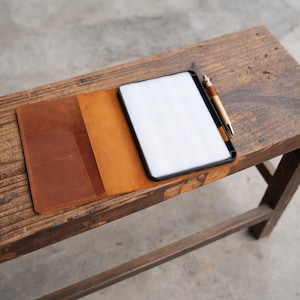 Puede incluir: Una funda de tableta de cuero marrón con una tableta y un bolígrafo. La funda está abierta, revelando un interior de color claro y dos bolsillos. El bolígrafo es marrón y plateado. Los artículos están sobre un banco de madera.