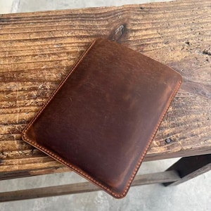 May include: A rectangular, brown leather case with a stitched border. The case has a distressed appearance, with variations in color and texture. The leather appears to be thick and durable, suggesting a protective function. The case is likely designed to hold a tablet or similar device.