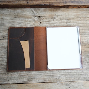May include: A brown leather notebook cover featuring a dark brown interior pocket with a tan accent strip. The right side holds a clear plastic sleeve containing a white notepad and a silver pen secured in a side loop, creating a functional set.