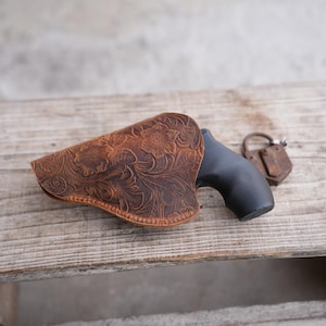 May include: A brown leather holster with a floral embossed design. The holster is positioned on a wooden surface, partially concealing a black handgun. An antique padlock rests nearby.