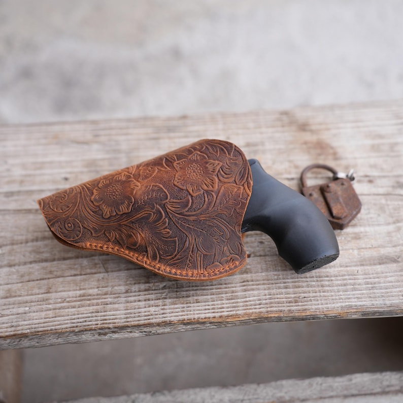 May include: A brown leather holster with a floral embossed design. The holster is positioned on a wooden surface, partially concealing a black handgun. An antique padlock rests nearby.