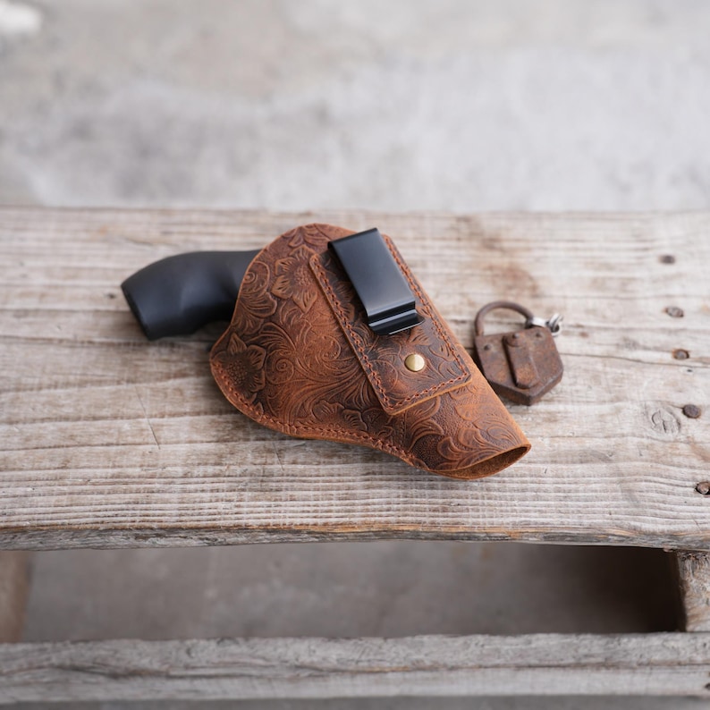 May include: A brown leather holster with a floral embossed design, a black clip, and a brass stud. A black handgun is partially visible inside the holster. An antique padlock rests beside it on a wooden surface.