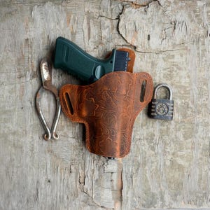 Puede incluir: Una funda de cuero marrón con un patrón floral, que contiene una pistola verde. También hay un candado plateado y unas tijeras oxidadas. Los objetos están dispuestos sobre una superficie de madera envejecida.