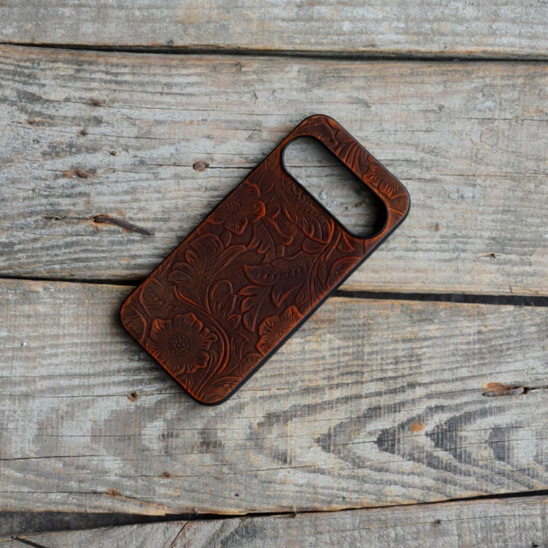 Puede incluir: Una funda de tel&eacute;fono de cuero marr&oacute;n con un dise&ntilde;o floral en relieve. La funda tiene un recorte para la lente de la c&aacute;mara.