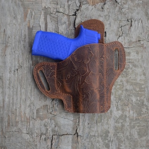 Puede incluir: Una funda de cuero marrón con un diseño floral en relieve, que sostiene una pistola azul. La funda tiene un aspecto desgastado y está sobre un fondo de madera envejecida.