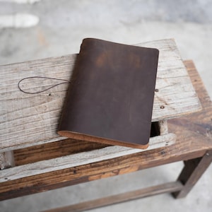 May include: A dark brown leather journal with a loop closure, resting on a rustic wooden bench. The journal's cover has a textured appearance, and the pages are visible along the edges. The bench is made of weathered wood.