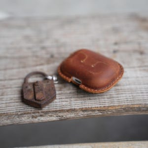 May include: A brown leather AirPod case with a stitched border and a small, rusty padlock. The case is rectangular and has a smooth, worn appearance. The padlock is small and has a keyhole.