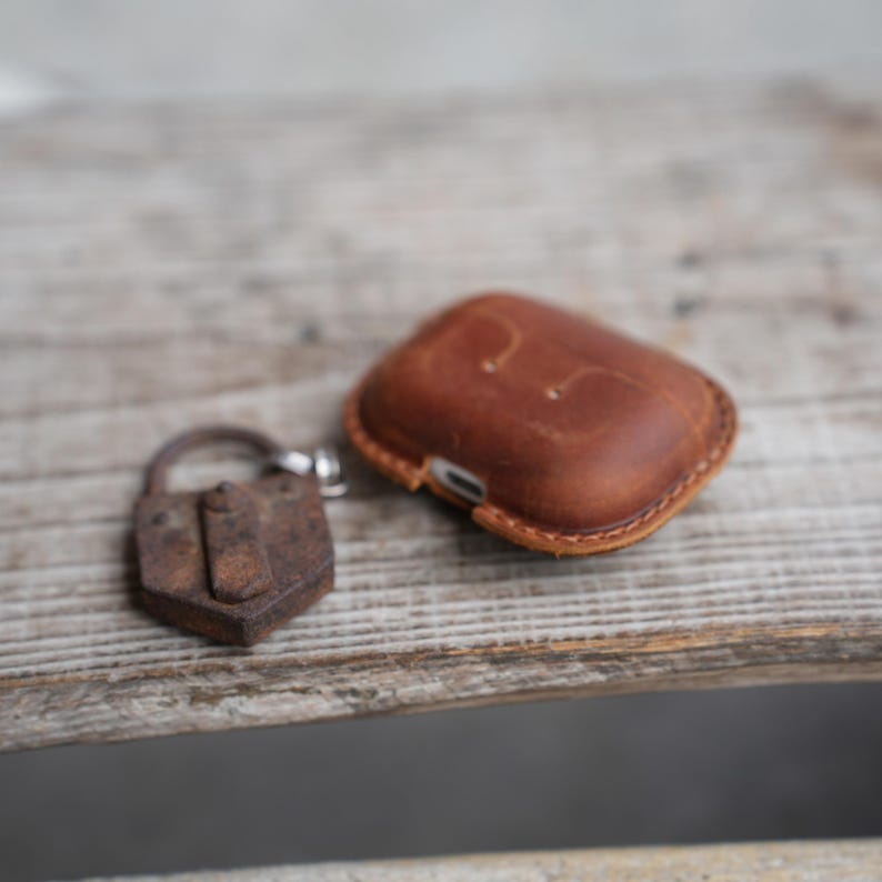 May include: A brown leather AirPod case with a stitched border and a small, rusty padlock. The case is rectangular and has a smooth, worn appearance. The padlock is small and has a keyhole.