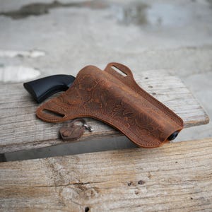 Puede incluir: Una funda de cuero marrón con un intrincado patrón floral grabado en la superficie. La funda contiene una pistola negra e incluye una pequeña etiqueta de cuero marrón a juego sujeta por una anilla metálica, con ranuras para cinturón visibles.