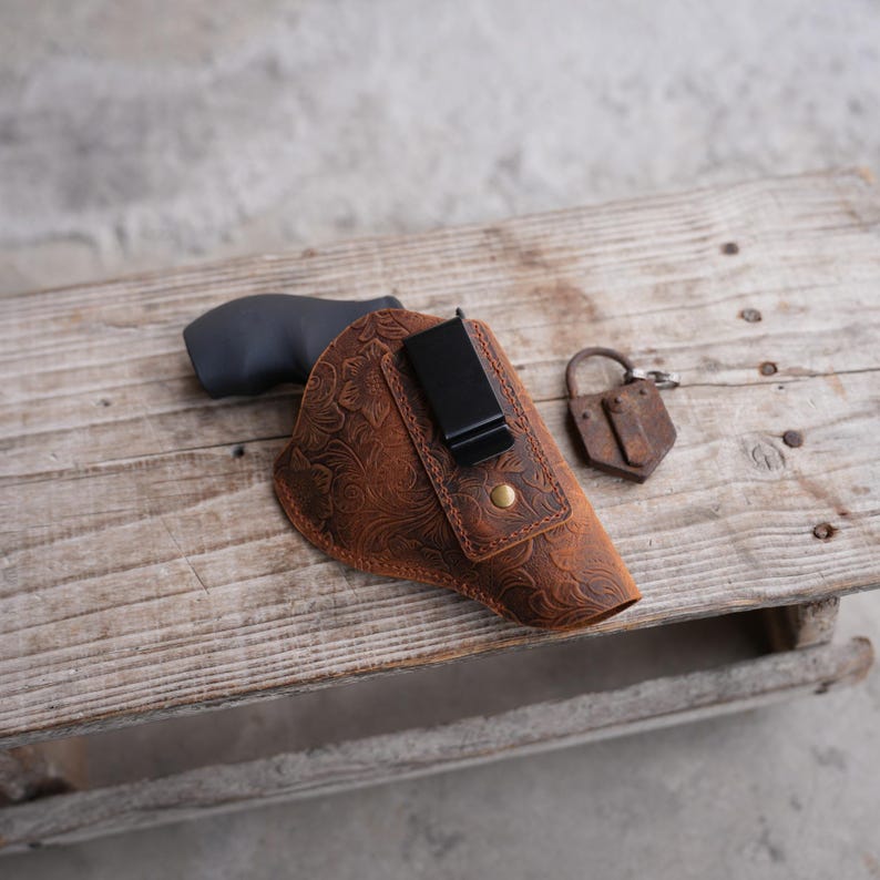 May include: A brown leather holster with a floral embossed design, a black clip, and a brass button. A black handgun is partially visible inside the holster. An antique padlock rests nearby. The items are on a wooden surface.