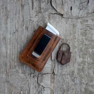 Może przedstawiać: Brązowy skórzany portfel z klips do pieniędzy i ozdobnym tłoczonym wzorem. Portfel leży na drewnianej powierzchni z rdzewiejącym zamkiem obok.