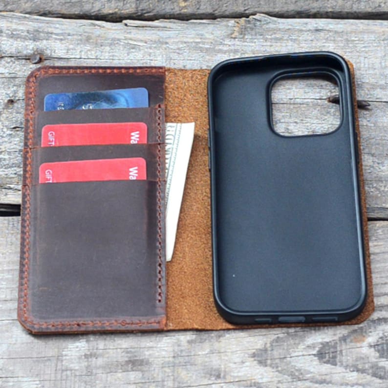 May include: A brown leather phone case with a built-in wallet. The case has slots for credit cards and cash. The phone case is open, revealing a black phone inside.
