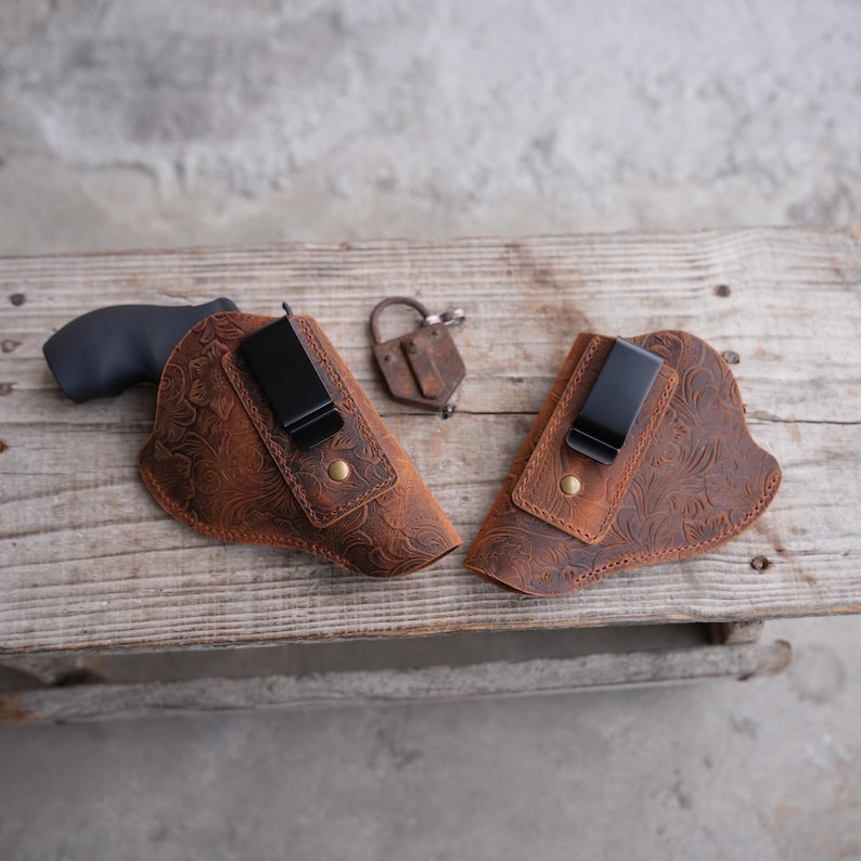 May include: Two brown leather gun holsters with floral embossing and black metal clips. A black revolver is in one holster. A small, brown leather pouch with a metal clasp is also present. The items are on a wooden surface.