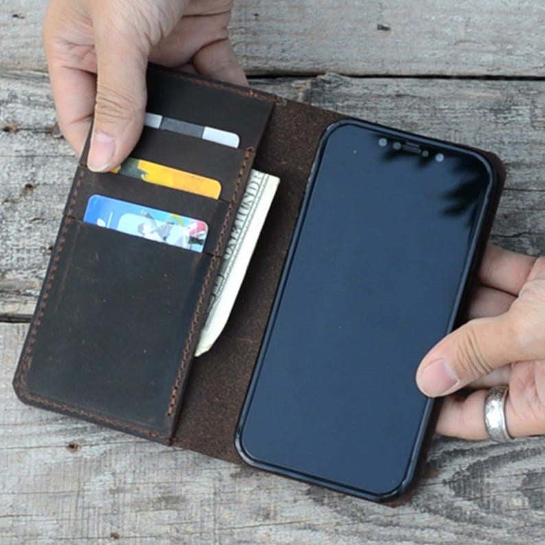May include: A brown leather phone case with a built-in card holder. The case is open, revealing a black smartphone and several cards inside the holder.