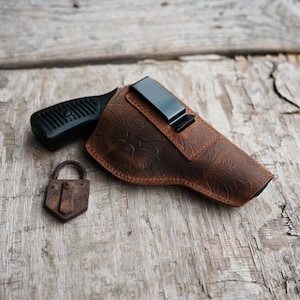 Op de afbeelding: Een bruin leren holster met een bloemmotief en een zwarte clip, naast een zwart pistool en een klein, roestig hangslot. De holster is ontworpen om het pistool te bevatten. De items liggen op een verweerd houten oppervlak.